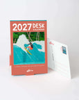 Small standing desk calendar for 2027 on a desk, next to an art card showing its postcard back
