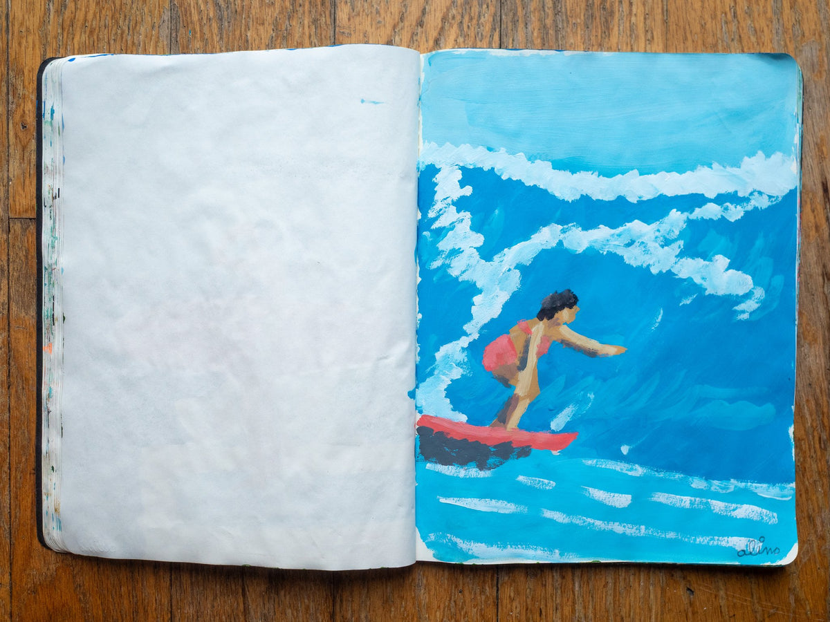 Drop 07 sketch 08 by Alimo in the open Moleskine sketchbook, painted on the right page only: a surfer in a pink swimsuit riding a curling blue wave